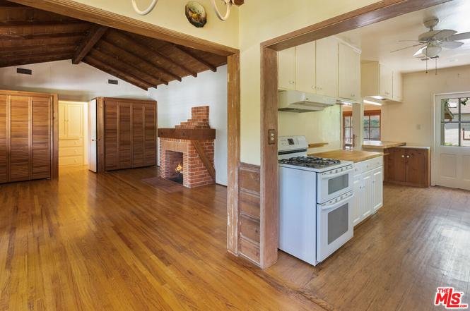 Kitchen with white appliances and wood cabinets open to living room with brick fireplace and wood beams