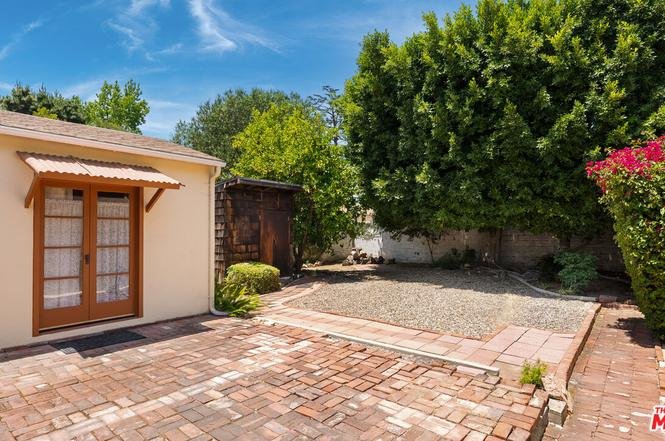 Backyard with brick patio, small building with brown doors, trees, and green plants