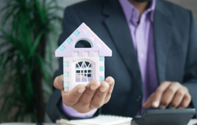 Person in suit holding a small house model and using a calculator