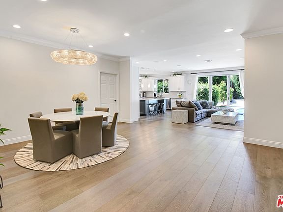 Dining area with round table, gray chairs, rug, plants, bench, and windows.