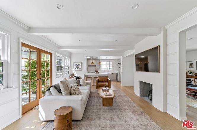 Modern living room with white sofa, pillows, wooden coffee table, large windows, and open kitchen.