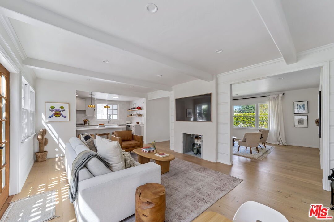 Modern living room with white sofa, wooden coffee table, TV, large windows, and open kitchen.