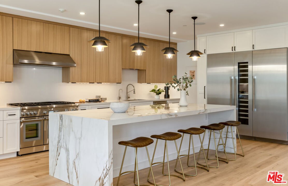 Kitchen with light wood cabinets, large island with white countertop, bar stools, stainless steel appliances, and pendant lights