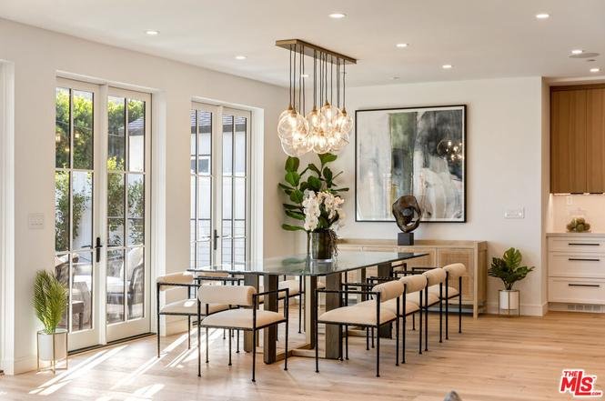 Dining area with glass table, beige chairs, big windows, plants, abstract wall art, and hanging light.