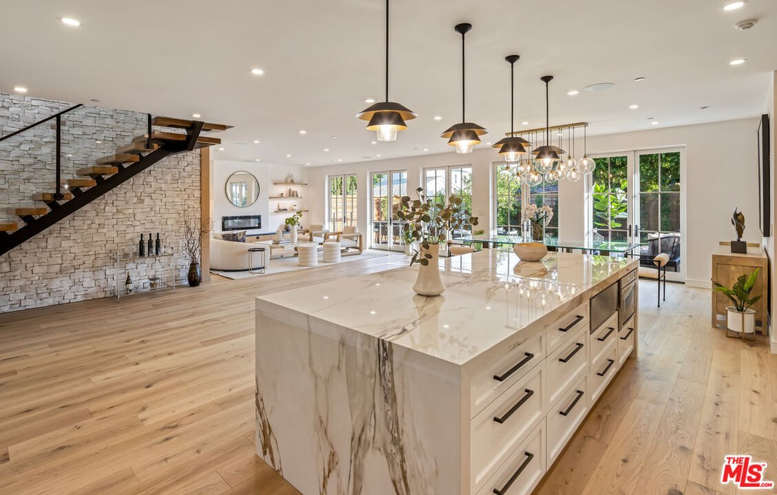 Kitchen with large marble island, pendant lights, open layout to living area, big windows, wood floor, and stairs to upper level.