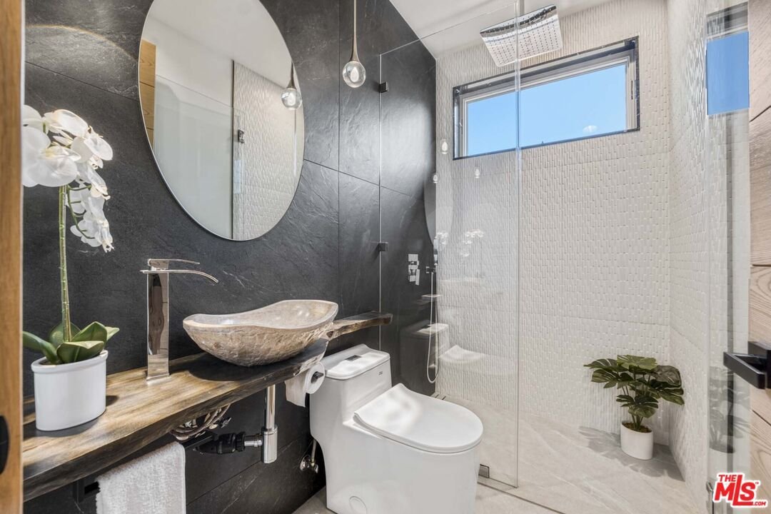 Bathroom with round mirror, bowl sink on wood counter, white toilet, glass shower, and small plants.