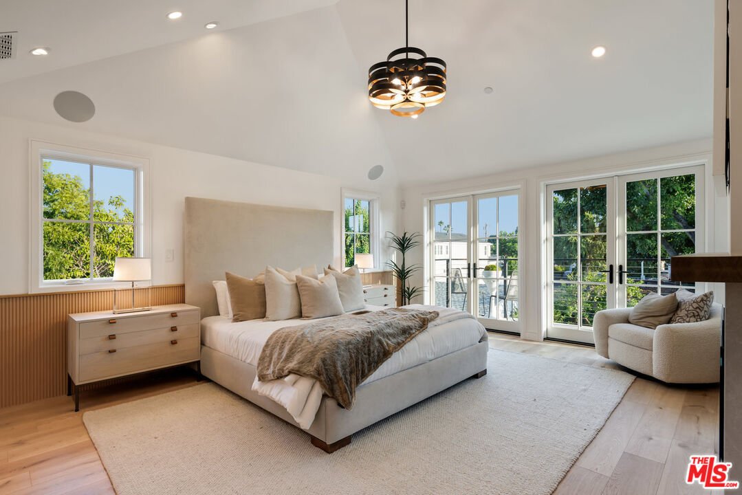 Bedroom with large bed, beige headboard, pillows, throw blanket, bedside lamps, armchair, wood floor, and big windows.