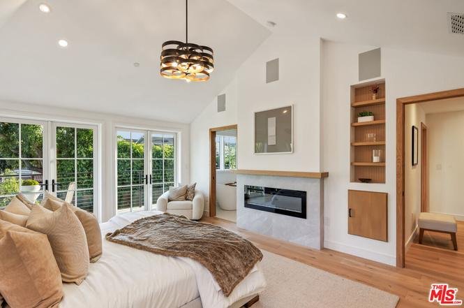 Bedroom with large bed, pillows, throw blanket, fireplace, big windows, and simple furniture.
