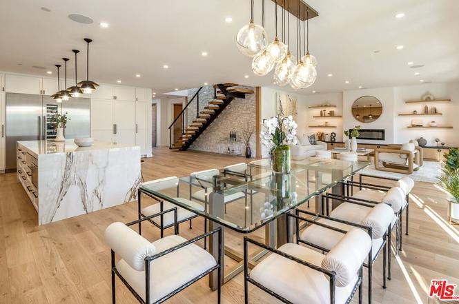 Dining area with glass table, soft chairs, kitchen in the back, and stairs nearby.