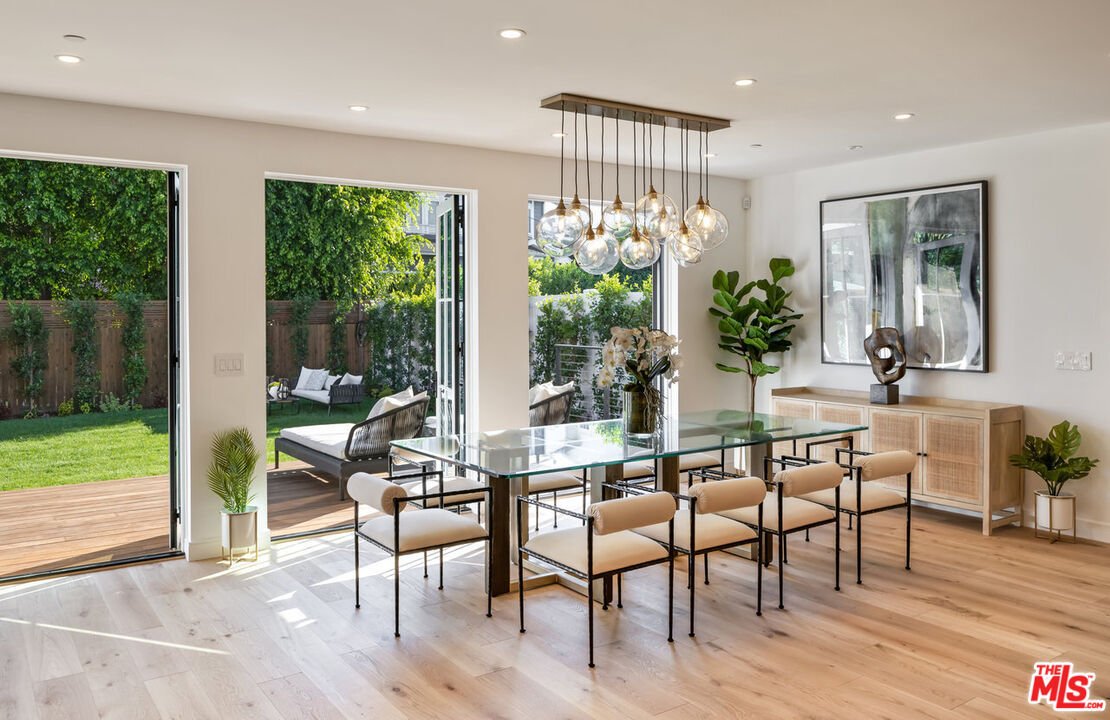 Dining room with glass table, eight chairs, large windows to backyard, and hanging ceiling lights.