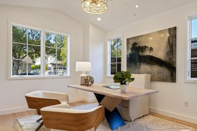 Home office with light desk, two wood chairs, lamp, plant, big windows, and abstract wall art.