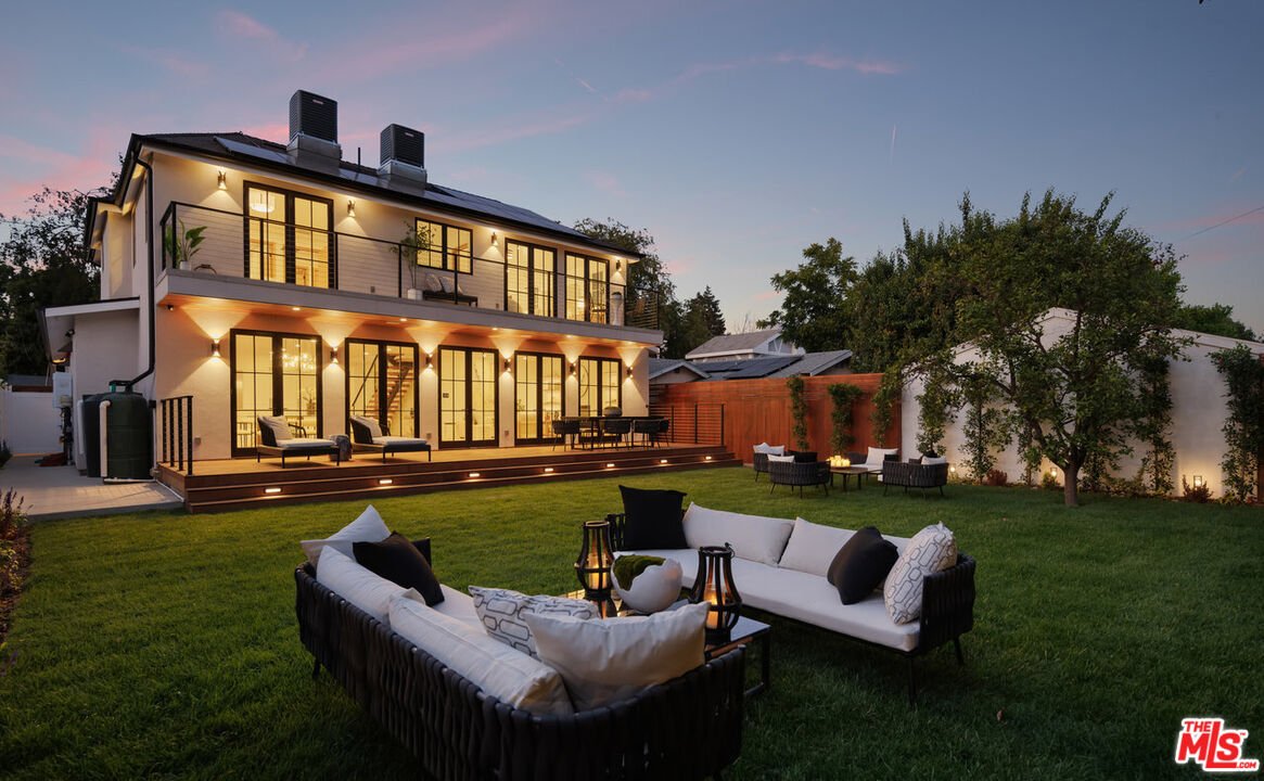 Backyard with green lawn, tall hedges, trees, and outdoor seating with wicker chairs and cushions.