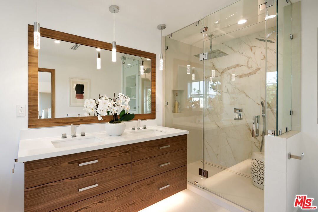 Bathroom with double sink vanity, big mirror, glass shower with marble wall, and white orchid plant.