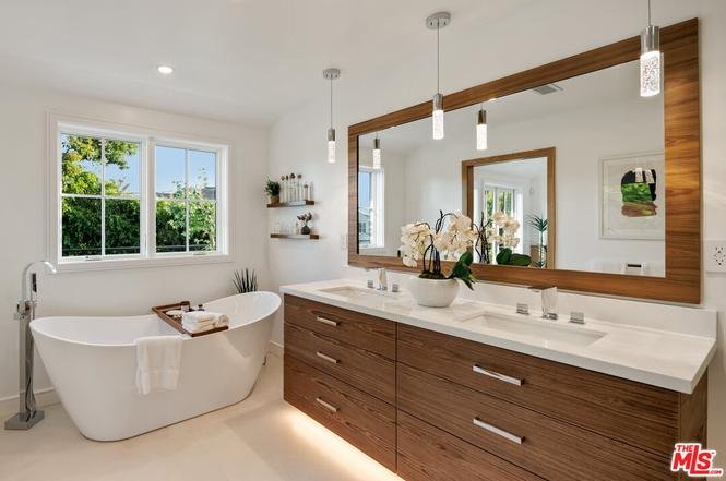 Bathroom with freestanding tub, double sink vanity, wood cabinets, big windows, and small plants.