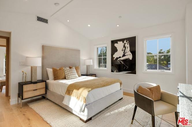 Bedroom with large bed, pillows, bedside lamps, desk with chair, wall art, and two windows.