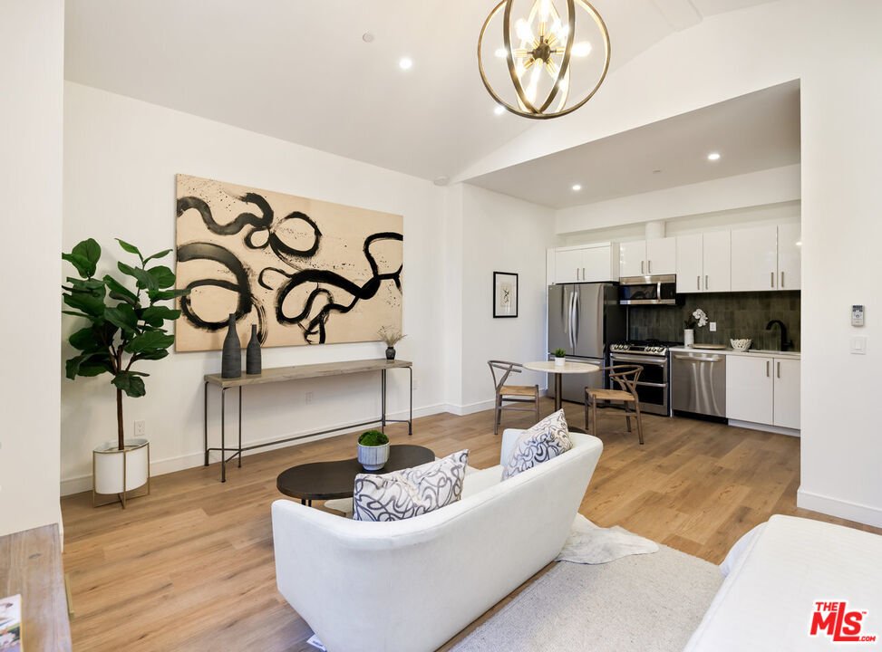 Living room with white couch, round coffee table, wall art, plant, dining chairs, and kitchen with steel appliances.