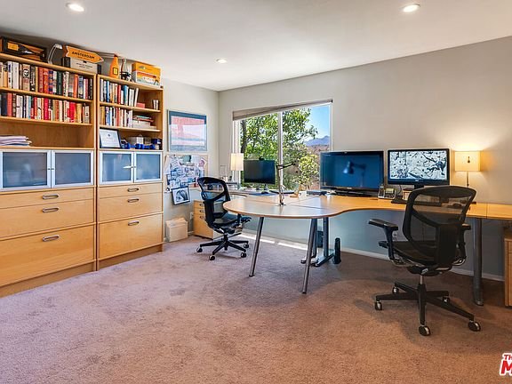 Home office with two desks, chairs, computer monitors, bookshelf with books, window with green view, and carpet floor.