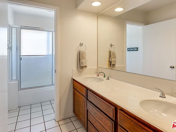 Bathroom with double sink vanity, wood cabinets, large mirrors, glass shower, and white tile floor.