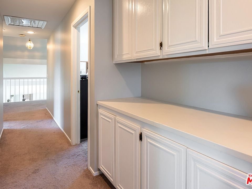Hallway with white cabinets, beige carpet, ceiling light, and doorway to another room.