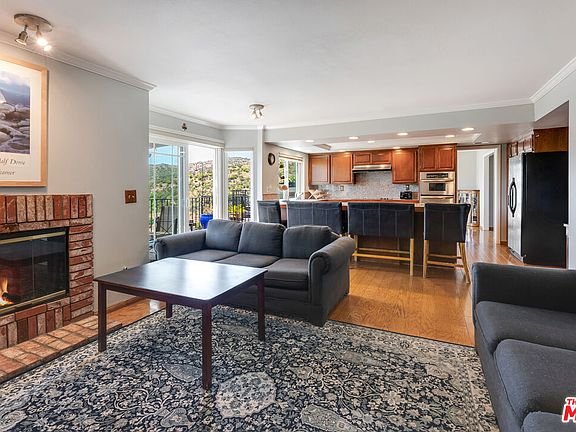 Living room with gray sofa, wood coffee table, fireplace, windows, rug, and kitchen with wood cabinets.