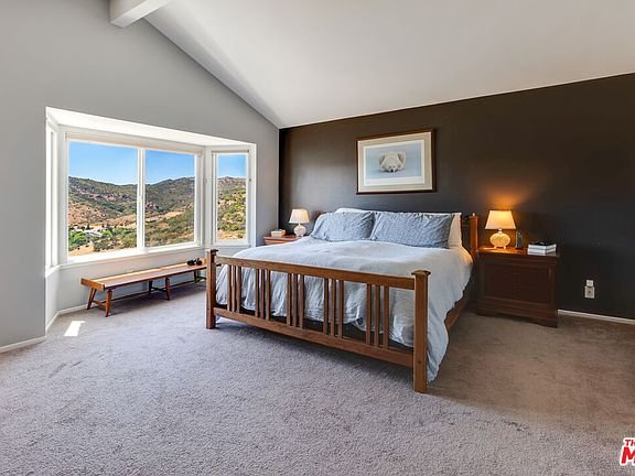 Bedroom with wood bed, blue bedding, bedside lamps, table, big window with hill view, and carpet floor.