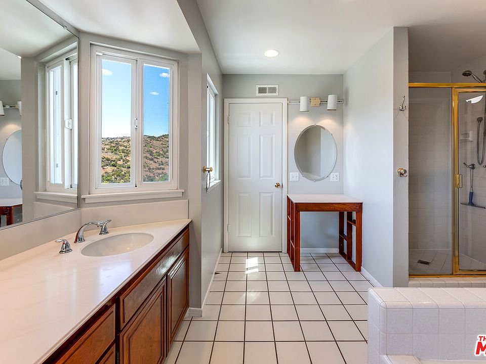 Bathroom with double sink vanity, large mirror, glass shower, bathtub, windows, and white tile floor.