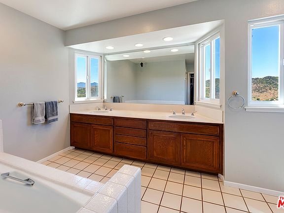Bathroom with double sink vanity, mirrors, bathtub, big windows, and beige tile floor.