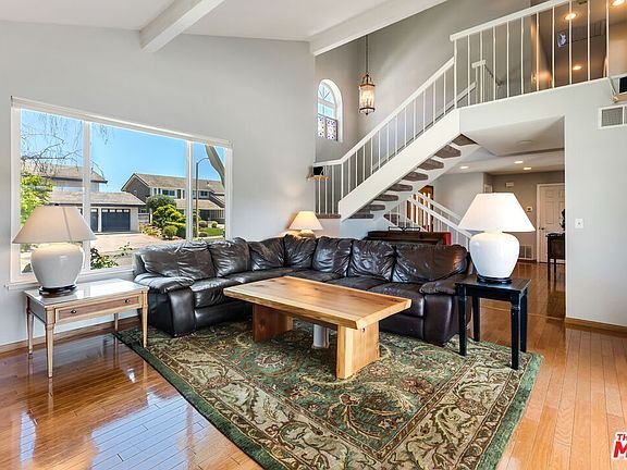 Living room with black leather sofa, wood coffee table, big windows, stairs in back, lamps, and wood floor.