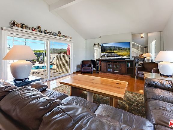Living room with leather sofa, wood coffee table, lamps, TV on cabinet, windows to pool view, plants, and wall art.