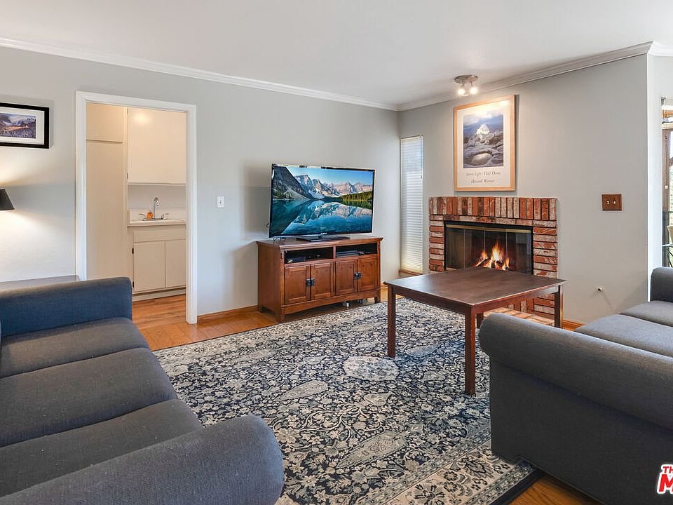 Living room with two gray sofas, wood coffee table, brick fireplace, TV, and kitchen through open doorway.