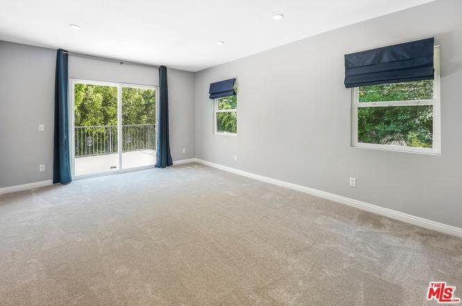 Room with gray walls, carpet floor, two windows with shades, and sliding glass door to balcony.