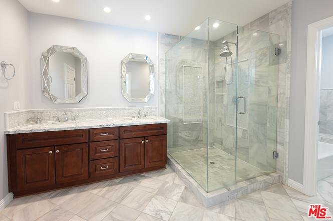 Bathroom with double sinks, wood cabinets, two mirrors, and glass shower with marble tiles.