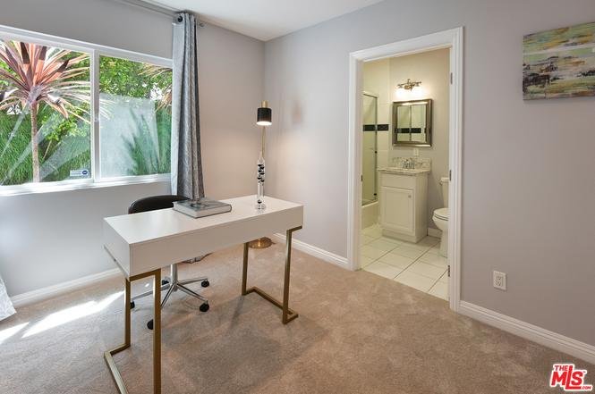 Office with desk, chair, window with garden view, bathroom door, gray walls, and carpet floor.
