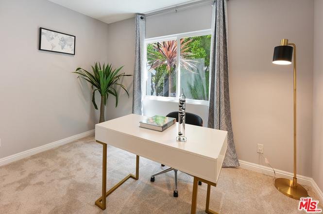 Home office with white desk, lamp, black chair, window with garden view, and plant.