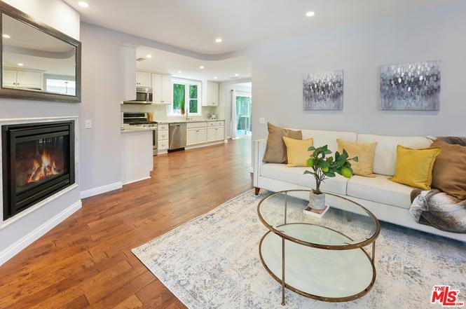 Living room with white sofa, pillows, glass coffee table, rug, fireplace, and open kitchen.