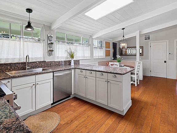Kitchen with white cabinets, granite counters, steel appliances, wood floor, big windows, and dining area in back.