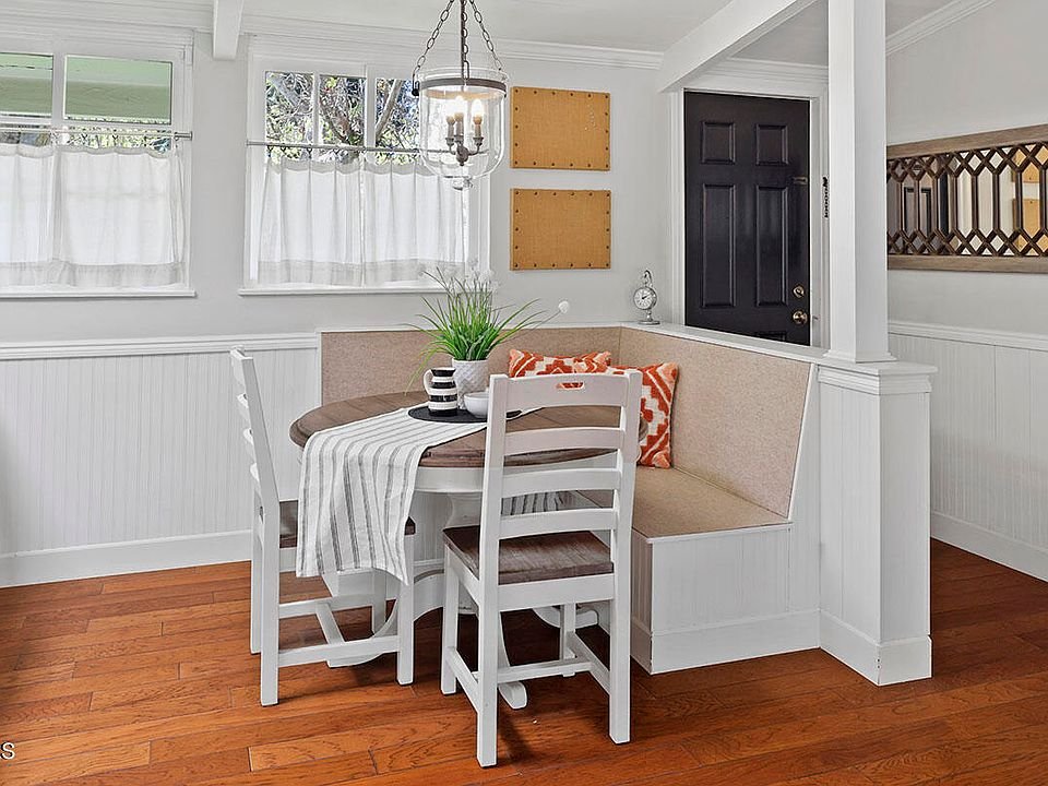 Dining nook with round table, two white chairs, bench seat, big windows, wall decor, and wood floor.