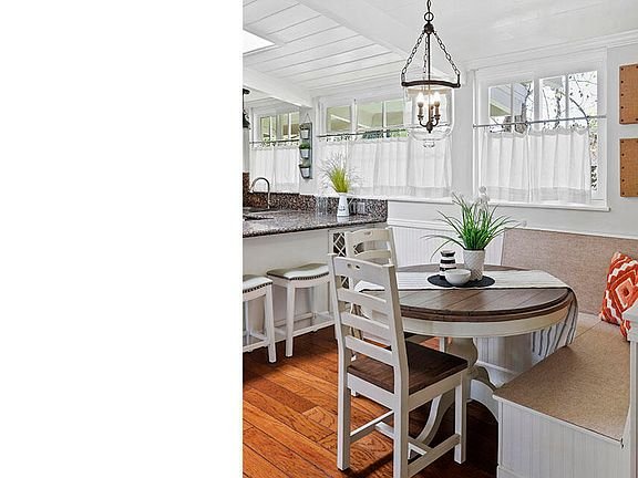Kitchen with round wood table, white chairs, bench with cushions, dark granite counter, plants, and big windows.