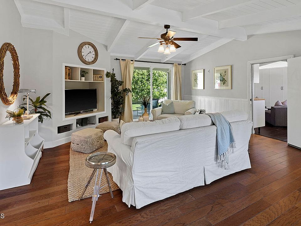 Living room with white sofa, wood floor, wall TV, plants, and big windows.