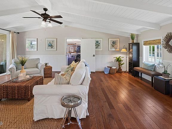 Living room with white sofa, wood floor, ceiling fan, plants, light walls, and high ceiling.