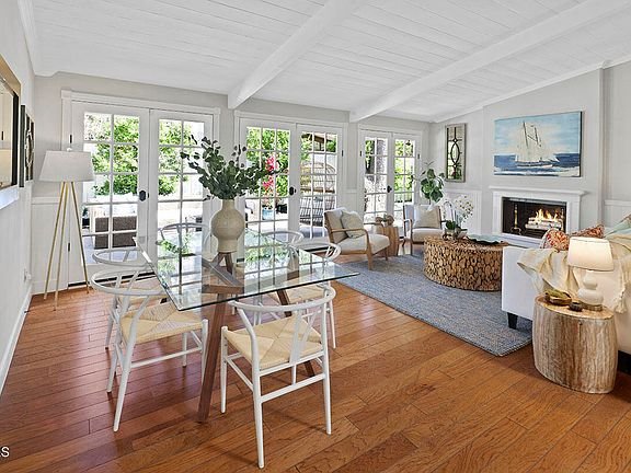 Living room with glass table, white chairs, fireplace, hardwood floor, and large windows.