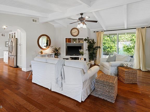 Living room with white sofa, wood floor, ceiling fan, big windows, TV on shelf, and plants.