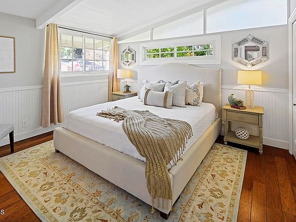 Bedroom with large bed, pillows, throw blanket, bedside lamps, big windows, wood floor, and rug.