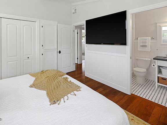Bedroom with white bedspread, wall TV, two closet doors, door to bathroom, and wood floor.