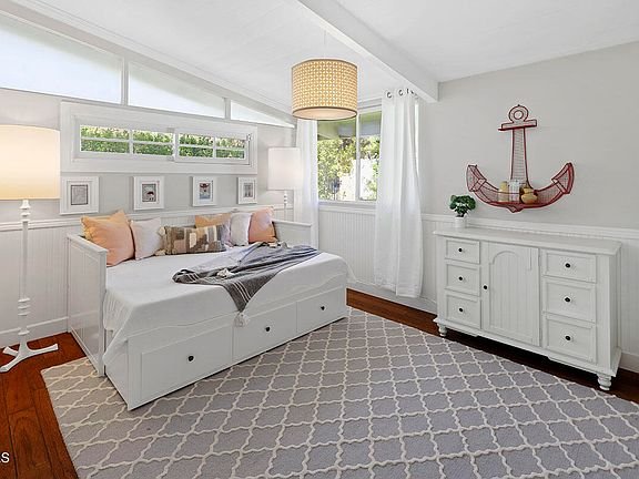 Bedroom with white bed with storage, pillows, large rug, dresser, wall decor, and big windows.