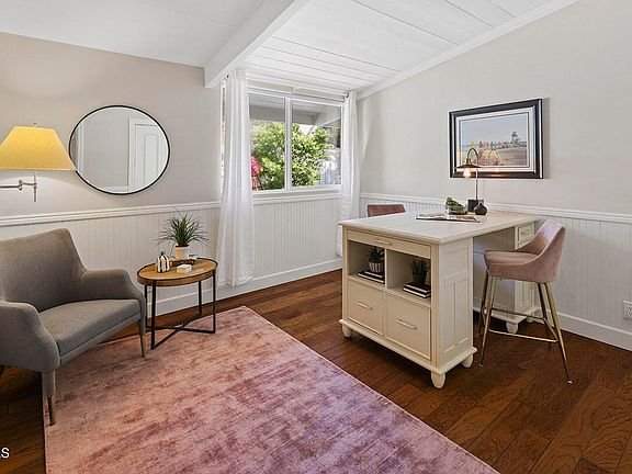 Home office with white desk, two pink chairs, round table with plant and cup, big windows, and wood floor.