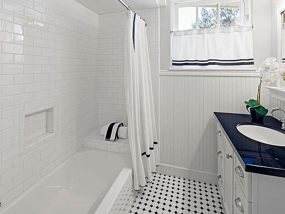 Bathroom with white tile shower, curtain, small shelf, dark counter, white cabinets, and black and white tile floor.