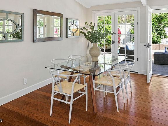 Dining room with glass table, white chairs, big windows, doors to patio, wall mirrors, and plant vase.