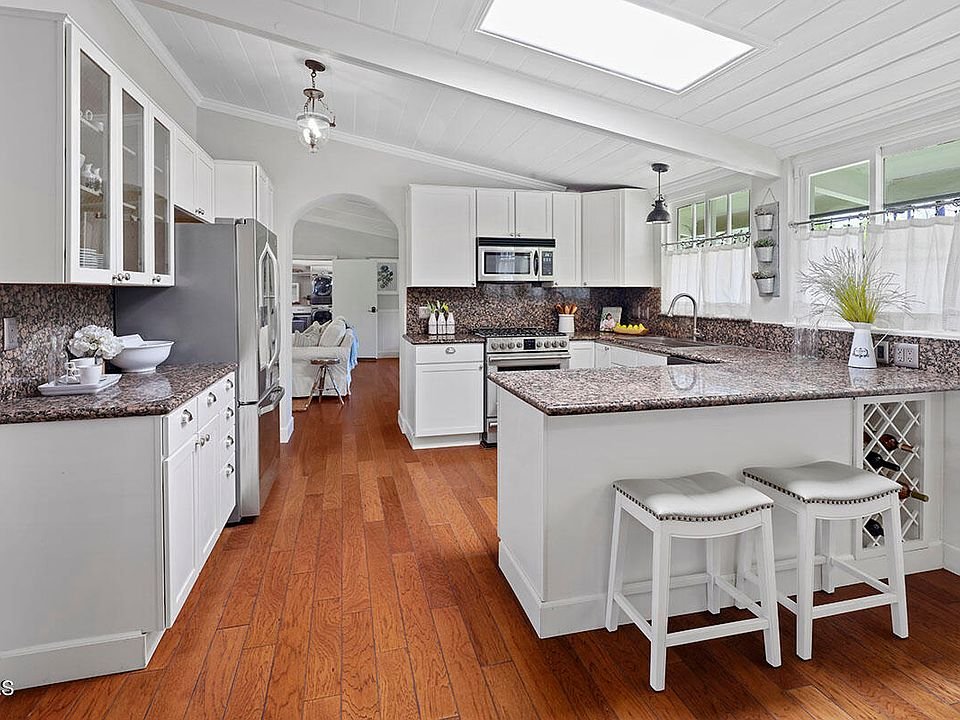 Kitchen with white cabinets, granite counters, steel appliances, island with bar stools, and wood floor.