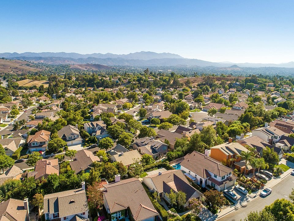 Aerial view of a suburban neighborhood with various houses surrounded by trees, hills, and mountains in the background under a clear blue sky.